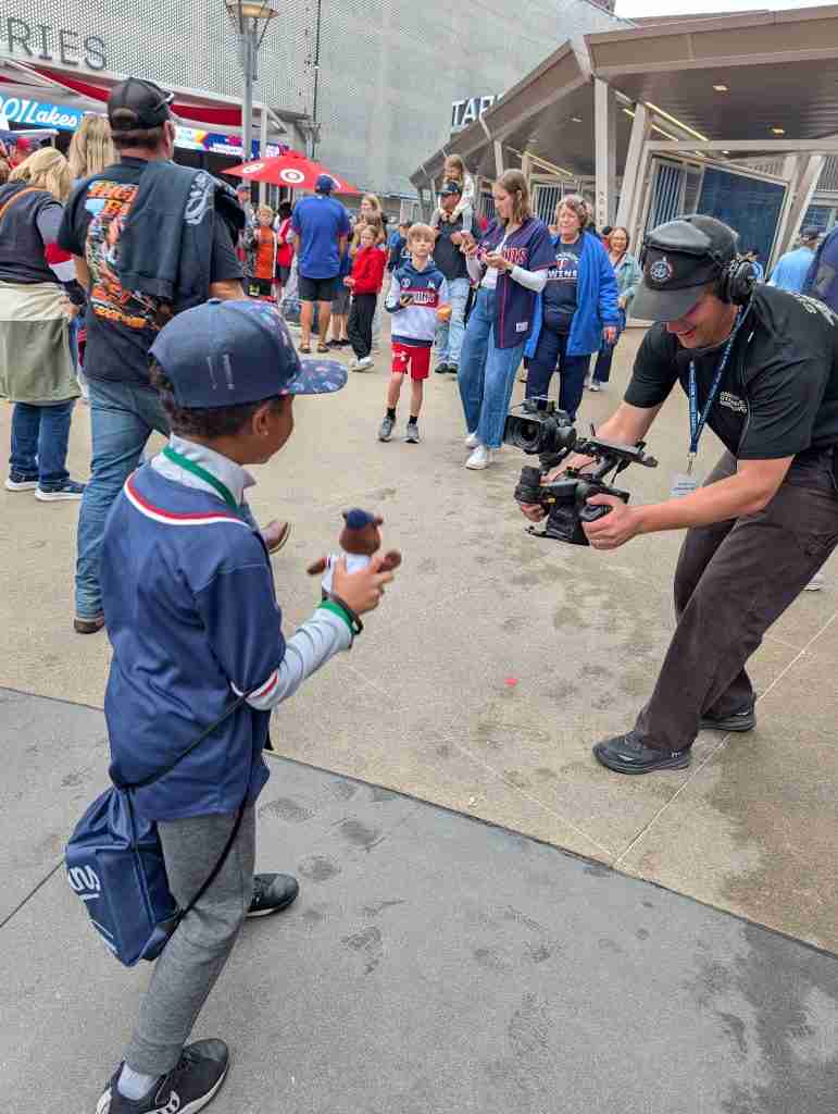Camera operator films a boy holding a T.C. Bear plush at Target Field